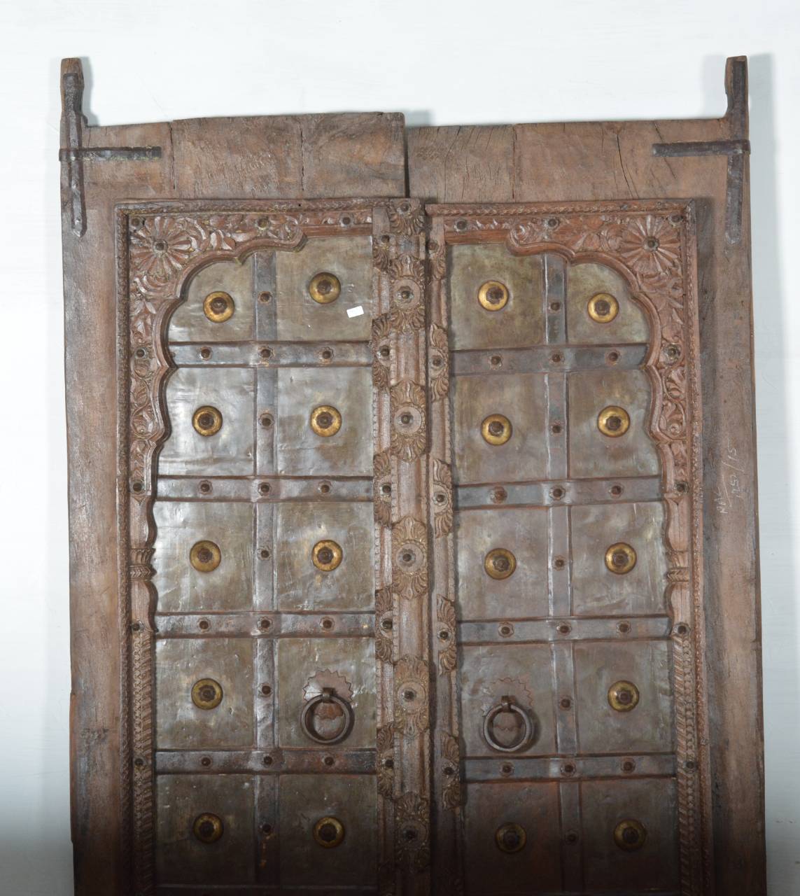 An antique carved wooden door with a weathered brown finish, featuring a grid of brass-toned metal studs and circular door knobs, photographed against a plain off-white background.