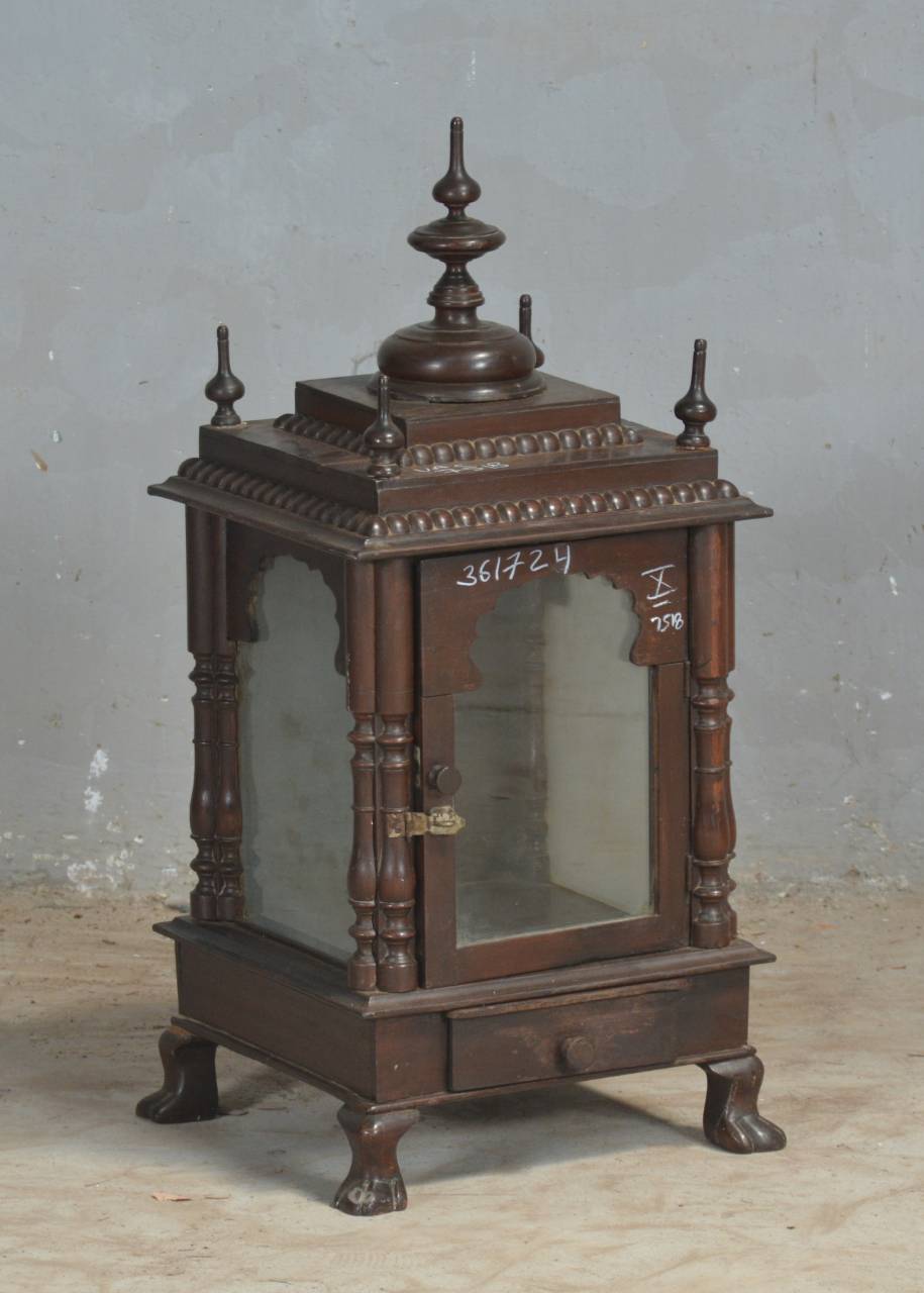 An ornate dark brown wooden display cabinet or miniature altar with glass doors, decorative finials, and carved legs, photographed against a plain gray wall.