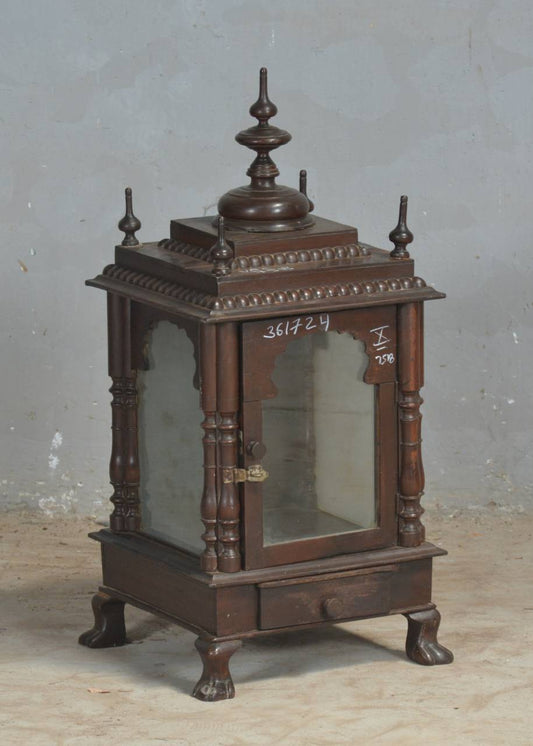 An ornate dark brown wooden display cabinet or miniature altar with glass doors, decorative finials, and carved legs, photographed against a plain gray wall.