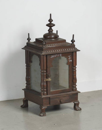 An ornate dark brown wooden display cabinet or miniature shrine with glass doors, decorative carved details, and a pointed finial on top, photographed against a plain light gray wall and floor.