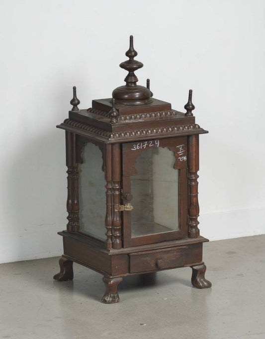 An ornate dark brown wooden display cabinet or miniature shrine with glass doors, decorative carved details, and a pointed finial on top, photographed against a plain light gray wall and floor.