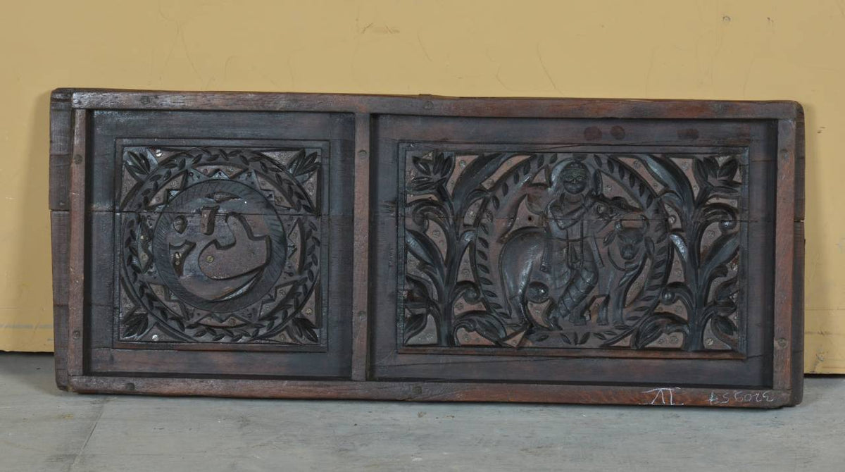 A dark carved wooden panel with two rectangular sections, featuring intricate relief carvings of a swan and a figure with a staff, set against a yellow wall and concrete floor.