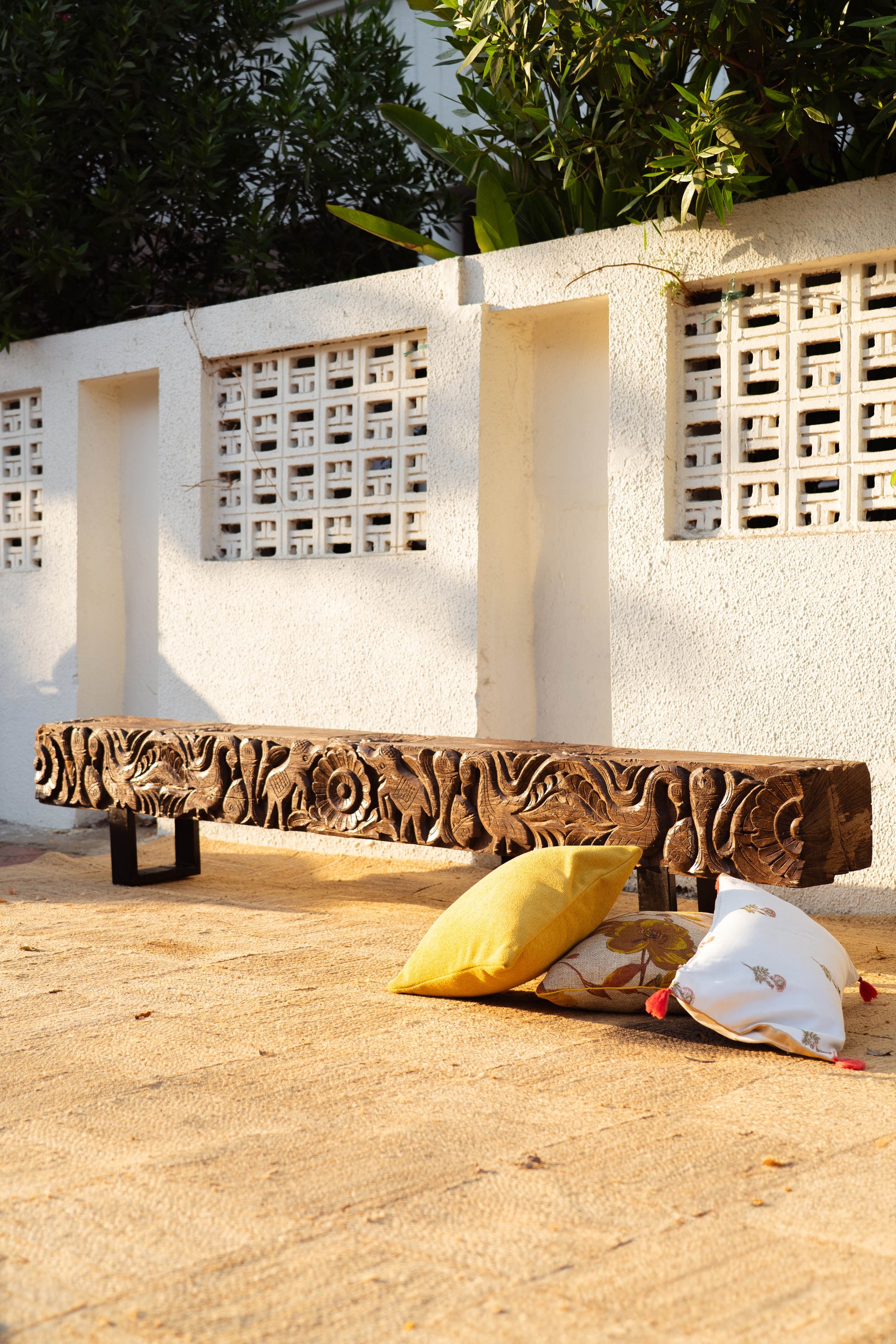 Carved wooden bench with decorative pillows on a sandy ground against a white wall.