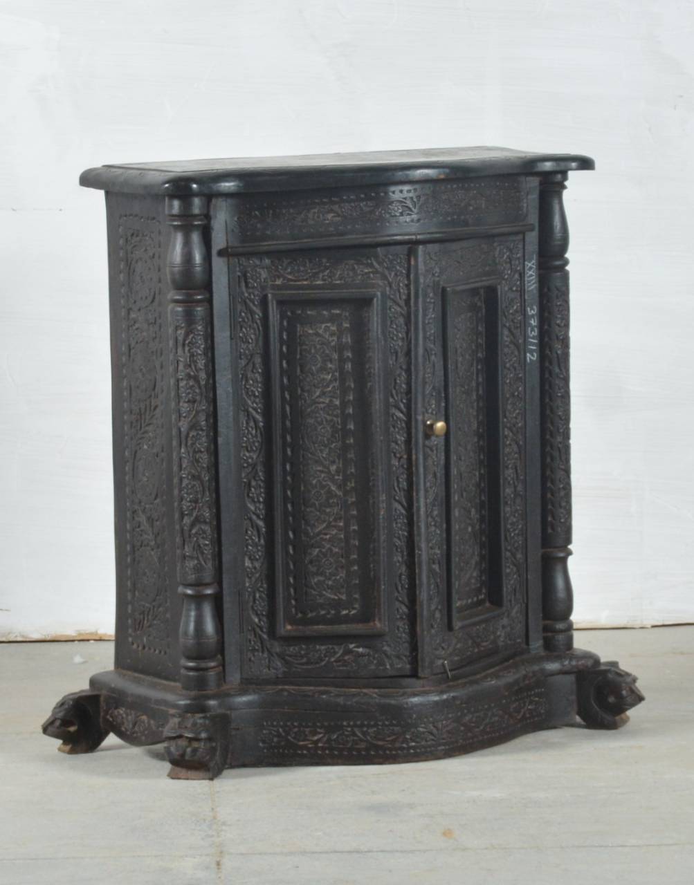 An antique carved wooden cabinet with two doors, ornate floral engravings, turned legs, and claw feet, finished in a dark black-brown tone with a small brass knob.