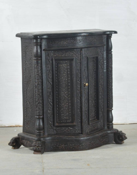 An antique carved wooden cabinet with two doors, ornate floral engravings, turned legs, and claw feet, finished in a dark black-brown tone with a small brass knob.