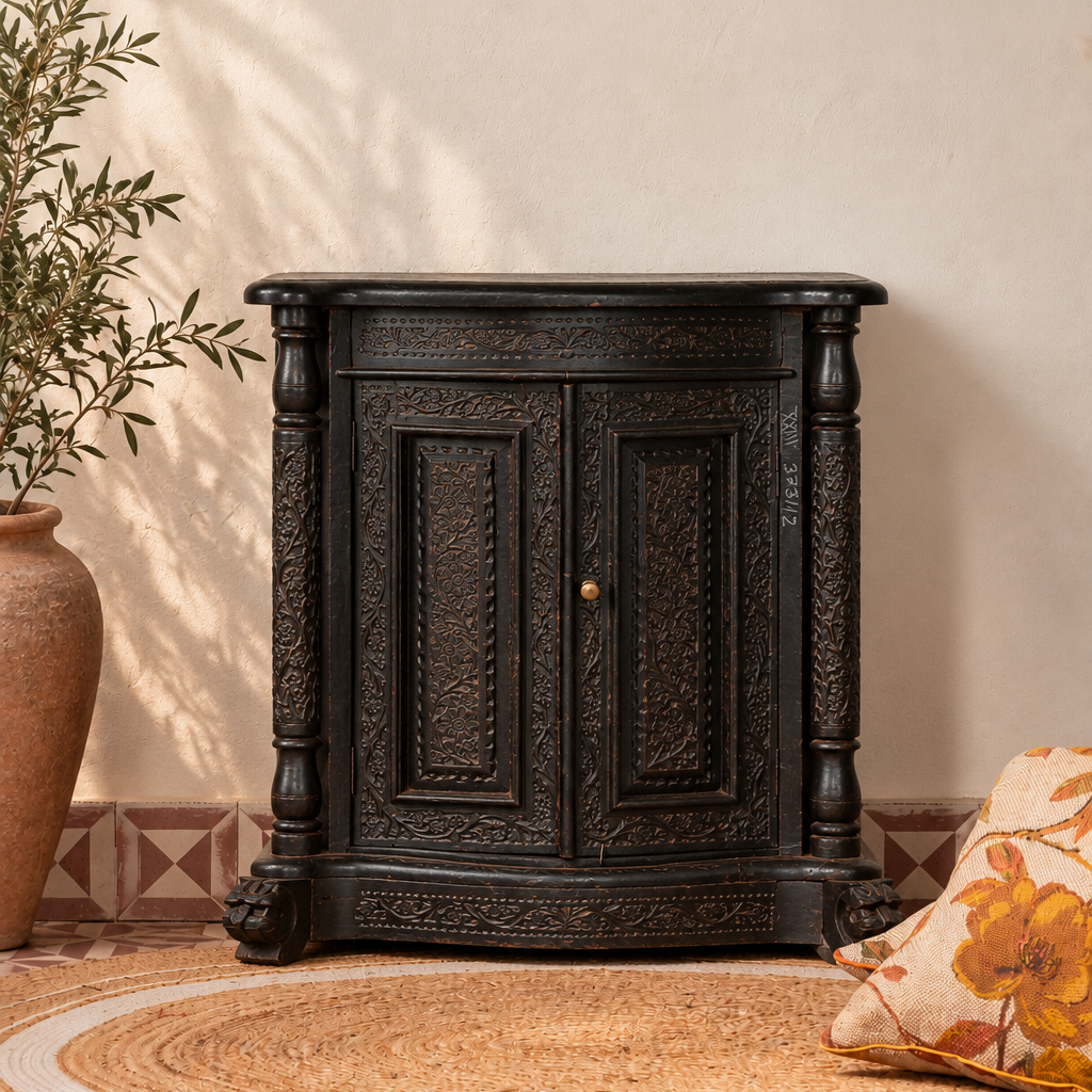 A dark carved wooden cabinet with ornate detailing and a small brass knob, styled in a warm room setting on a woven rug with a patterned tile floor, a potted plant, and a floral pillow.