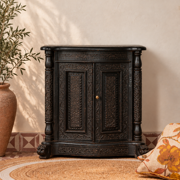 A dark carved wooden cabinet with ornate detailing and a small brass knob, styled in a warm room setting on a woven rug with a patterned tile floor, a potted plant, and a floral pillow.