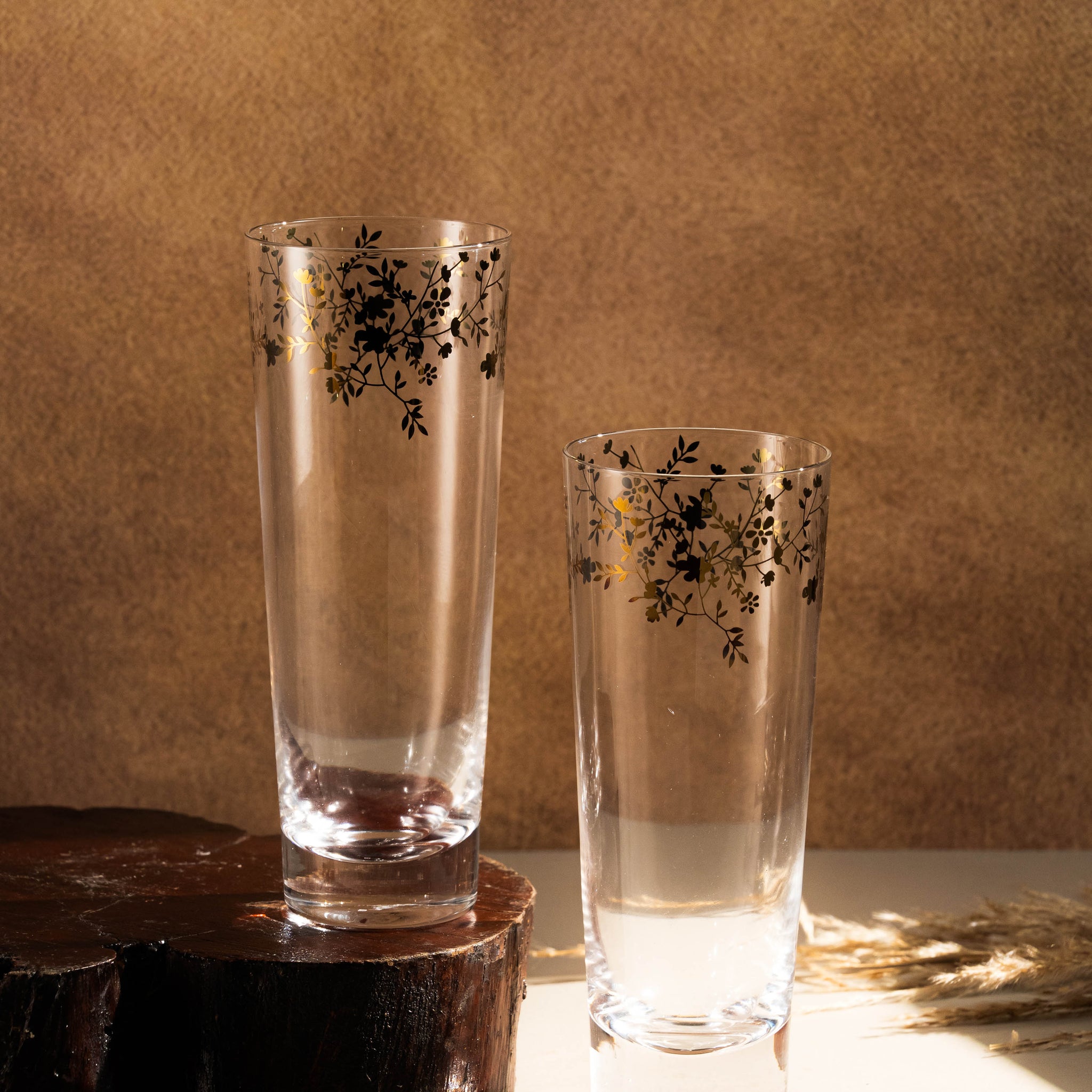 Two tall glasses with floral patterns on a wooden surface against a brown background