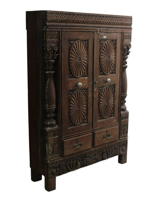 An antique dark brown carved wooden cabinet with ornate floral and geometric patterns, featuring two doors with circular sunburst carvings, two drawers at the bottom, and decorative side pillars.