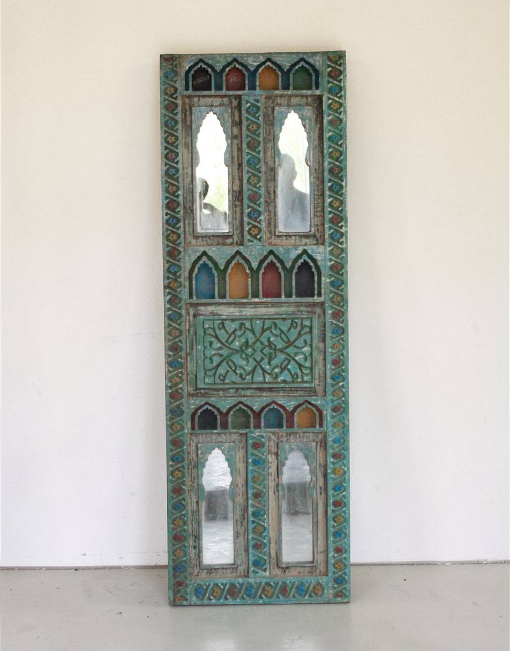 An ornate vertical decorative wall mirror panel with intricate turquoise, green, and gold geometric patterns and multiple arched mirror sections, leaning against a plain off-white wall.