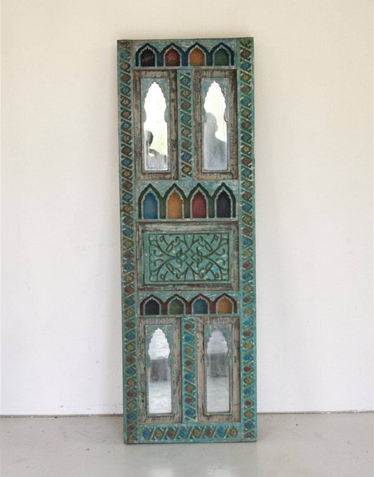 An ornate vertical decorative wall mirror panel with intricate turquoise, green, and gold geometric patterns and multiple arched mirror sections, leaning against a plain off-white wall.