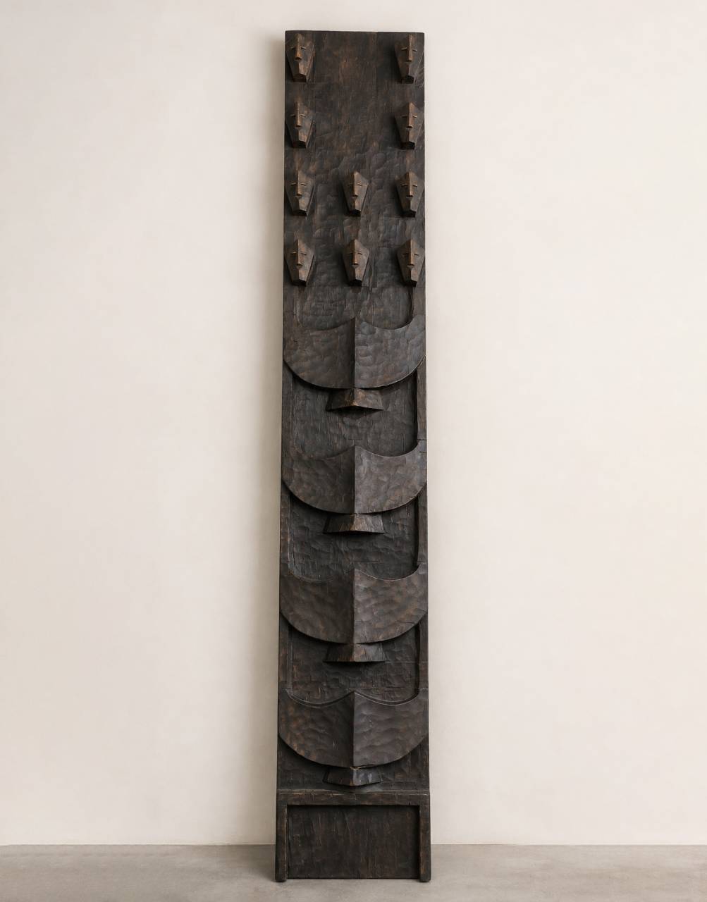 A tall, narrow dark carved wooden wall art piece with layered abstract leaf-like shapes and small hook-like protrusions near the top, photographed against a plain off-white wall and light gray floor.