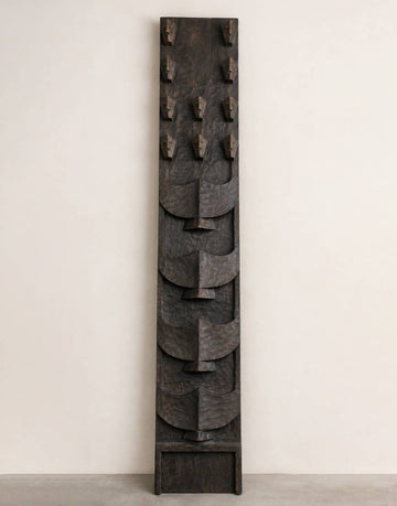 A tall, narrow dark carved wooden wall art piece with layered abstract leaf-like shapes and small hook-like protrusions near the top, photographed against a plain off-white wall and light gray floor.