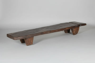 A rustic dark brown wooden bench made from a live-edge plank, supported by four thick block legs, set against a plain gray wall and concrete floor.