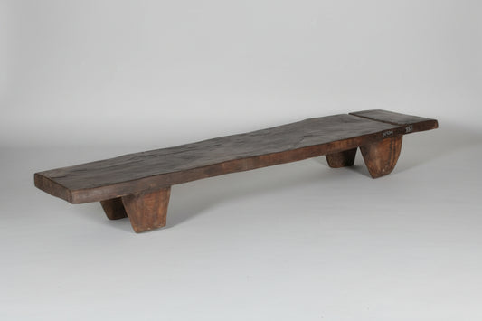 A rustic dark brown wooden bench made from a live-edge plank, supported by four thick block legs, set against a plain gray wall and concrete floor.