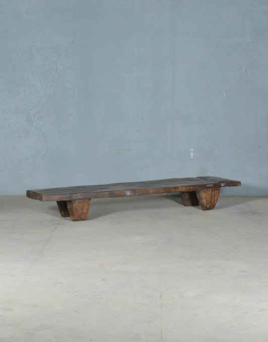 A rustic dark wood bench with a thick plank seat and four short block legs, photographed against a plain blue-gray wall and light concrete floor.