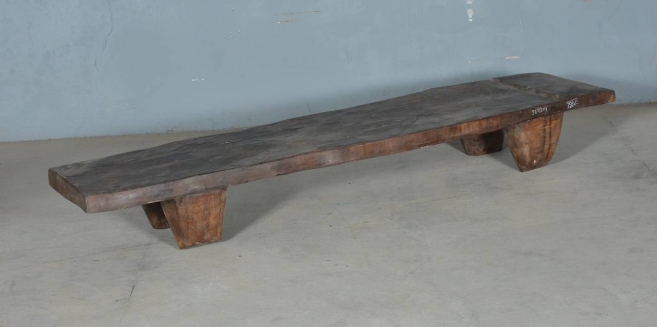A rustic dark brown wooden bench made from a live-edge plank, supported by four thick block legs, set against a plain gray wall and concrete floor.