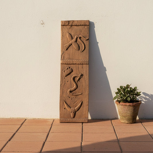A vertical rectangular carved wooden panel with three horizontal bands featuring embossed animal motifs, including a snake, a bird, and a horned figure, set against a plain off-white wall.