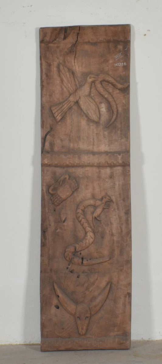 A tall, narrow carved wooden wall panel with relief carvings of birds, a snake, and a fish, shown against a plain off-white wall.