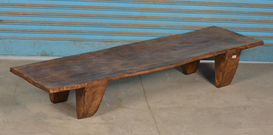 A rustic live-edge wooden bench with thick, tapered legs, shown against a blue corrugated metal wall and concrete floor.