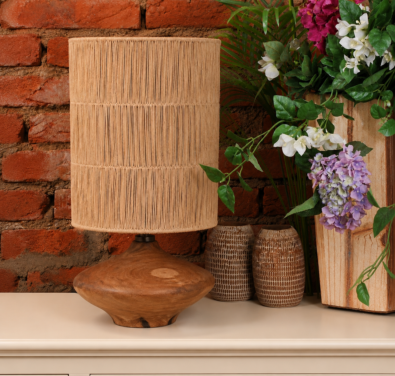 A rustic table lamp with a natural wood base and a cylindrical woven reed or straw shade, styled on a cream-colored surface against a red brick wall with flowers and wicker vases.