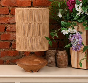 A rustic table lamp with a natural wood base and a cylindrical woven reed or straw shade, styled on a cream-colored surface against a red brick wall with flowers and wicker vases.