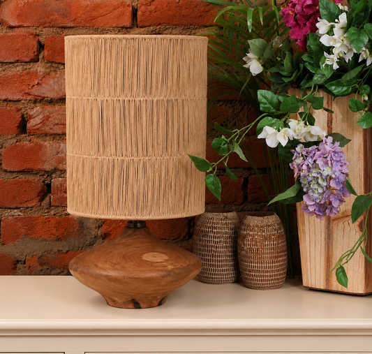 A rustic table lamp with a natural wood base and a cylindrical woven reed or straw shade, styled on a cream-colored surface against a red brick wall with flowers and wicker vases.