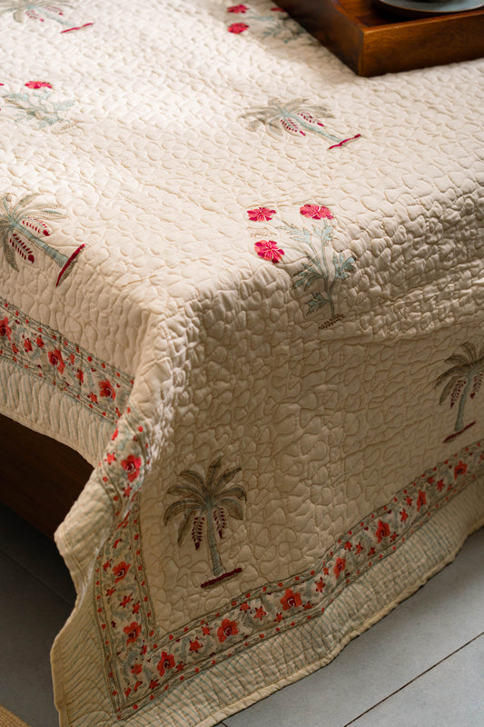 Beige quilt with floral and palm tree patterns on a wooden surface