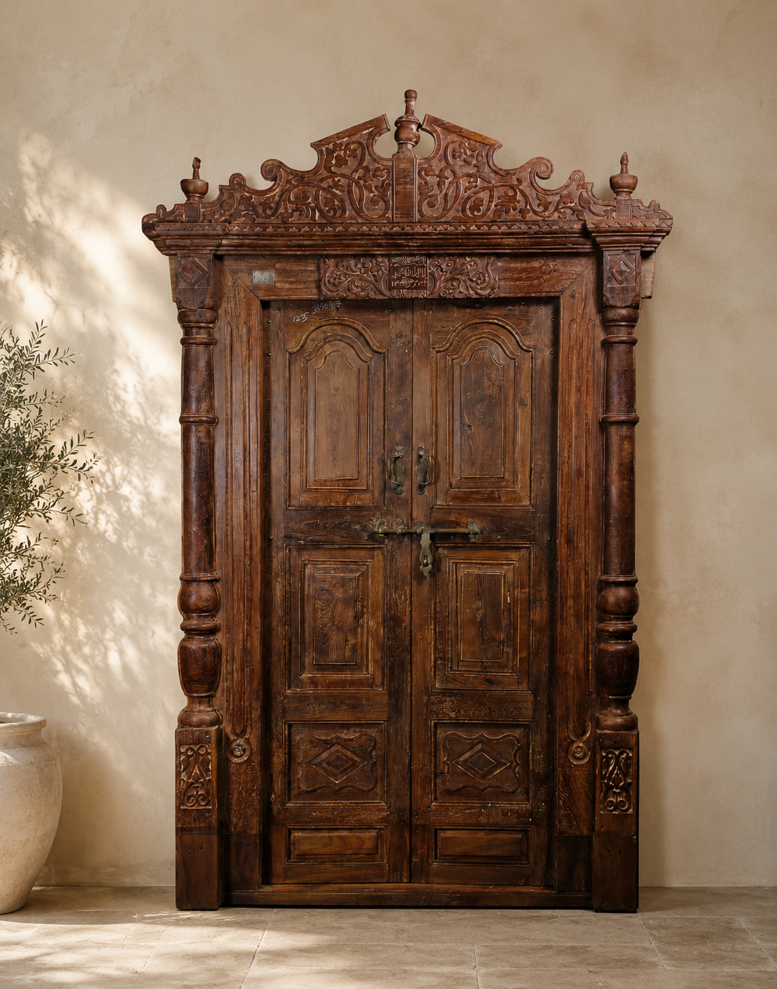 An ornate dark brown carved wooden double door with intricate relief detailing, set against a textured beige plaster wall with soft sunlight shadows.