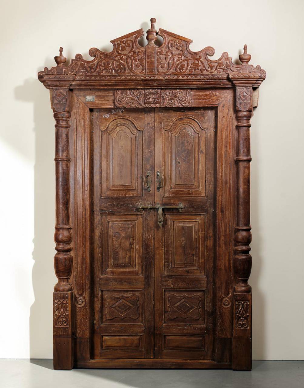 An ornate dark brown carved wooden double door with intricate relief details, traditional metal latch closures, and decorative turned side pillars, set against a plain off-white wall.