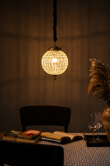 A warm glowing spherical pendant light with a textured glass or crystal-like shade hangs above a table in a dimly lit interior. A dark chair, books, an open book, a wine glass, and dried pampas grass in a vase are visible in the room.