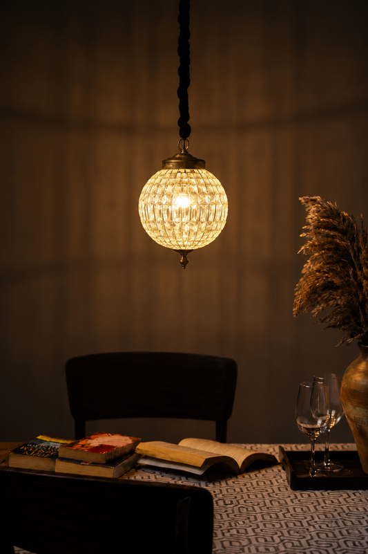 A warm glowing spherical pendant light with a textured glass or crystal-like shade hangs above a table in a dimly lit interior. A dark chair, books, an open book, a wine glass, and dried pampas grass in a vase are visible in the room.