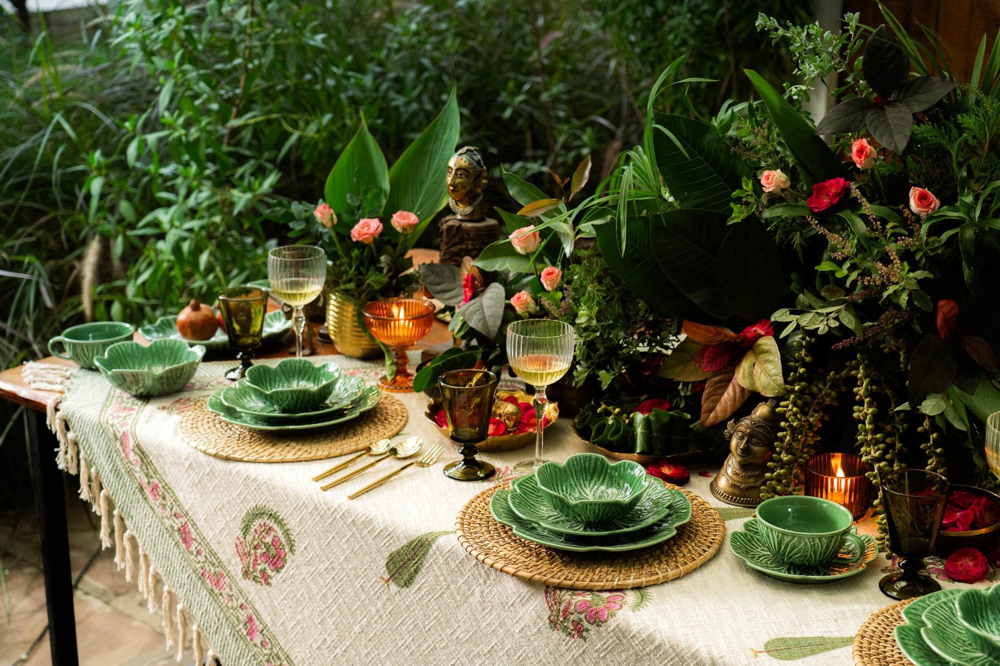 Dining table set with green plates, glasses, and a floral tablecloth in an outdoor setting with plants and candles.