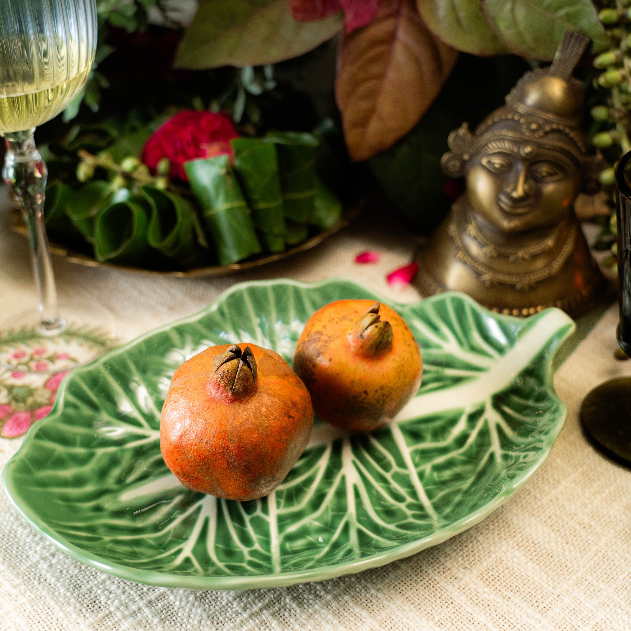 Two pomegranates on a decorative leaf-shaped plate with a glass of white wine and floral background.