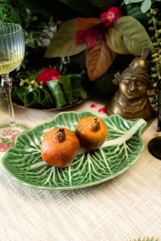 Two pomegranates on a decorative leaf-shaped plate with a glass of white wine and floral background.