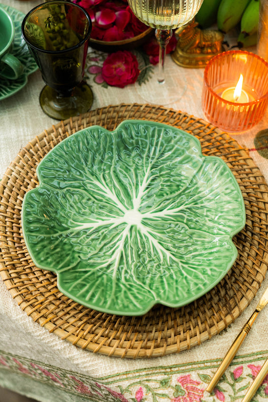 Green leaf-shaped ceramic plate on a woven placemat with candles and flowers in the background.
