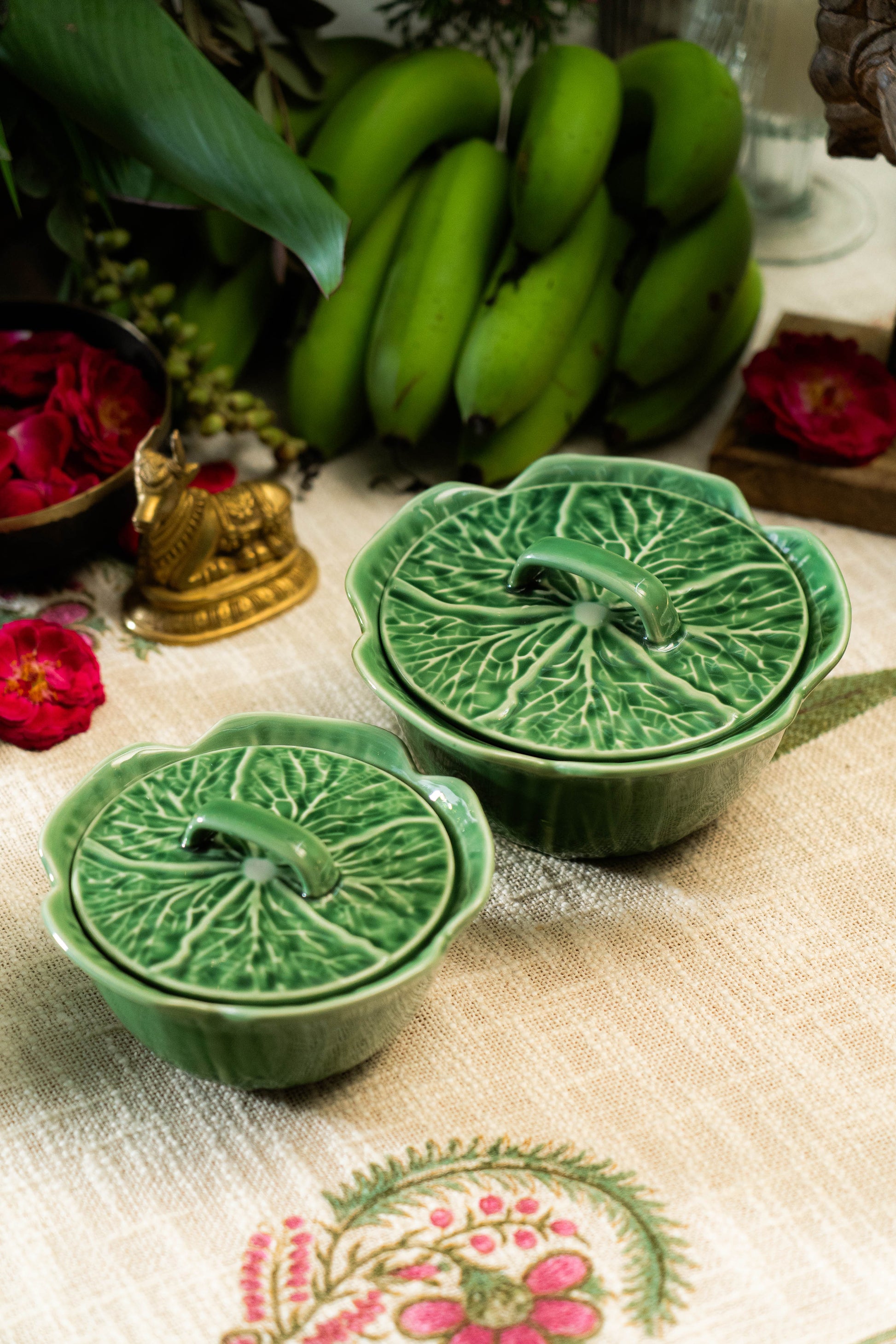 Two green ceramic pots with leaf patterns on a decorative table with flowers and bananas.
