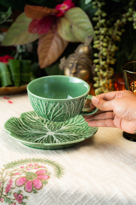 Green ceramic cup and saucer on a floral-patterned tablecloth with a hand holding the cup.