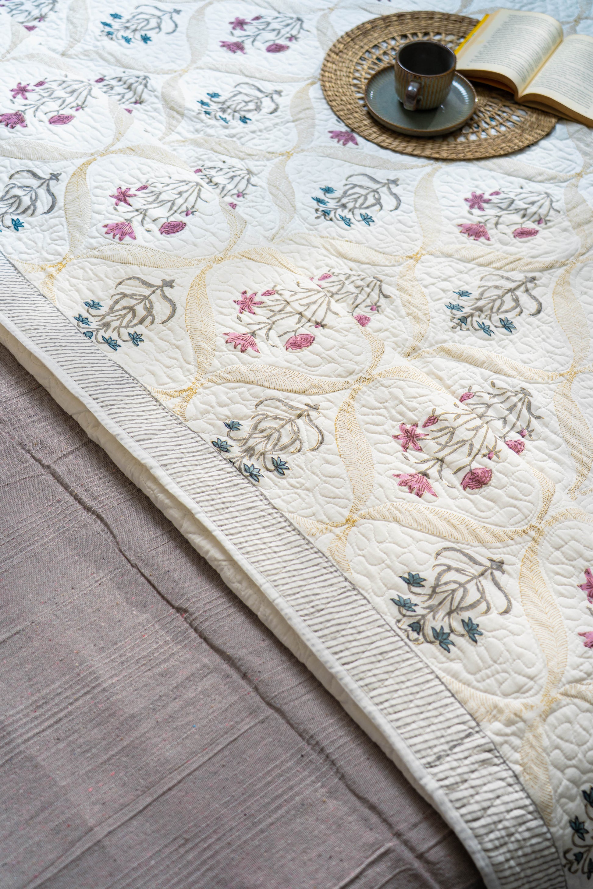 Floral-patterned quilt on a bed with a cup and book on a wooden table.