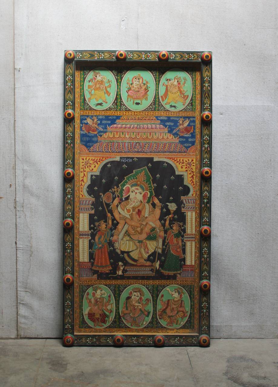 A large, hand-painted wooden religious panel featuring a central Hindu deity seated on a throne, surrounded by smaller oval and rectangular illustrations, with ornate carved corners and decorative borders, photographed against a gray concrete wall.