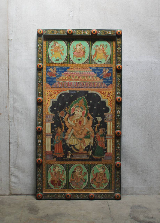 A large, hand-painted wooden religious panel featuring a central Hindu deity seated on a throne, surrounded by smaller oval and rectangular illustrations, with ornate carved corners and decorative borders, photographed against a gray concrete wall.
