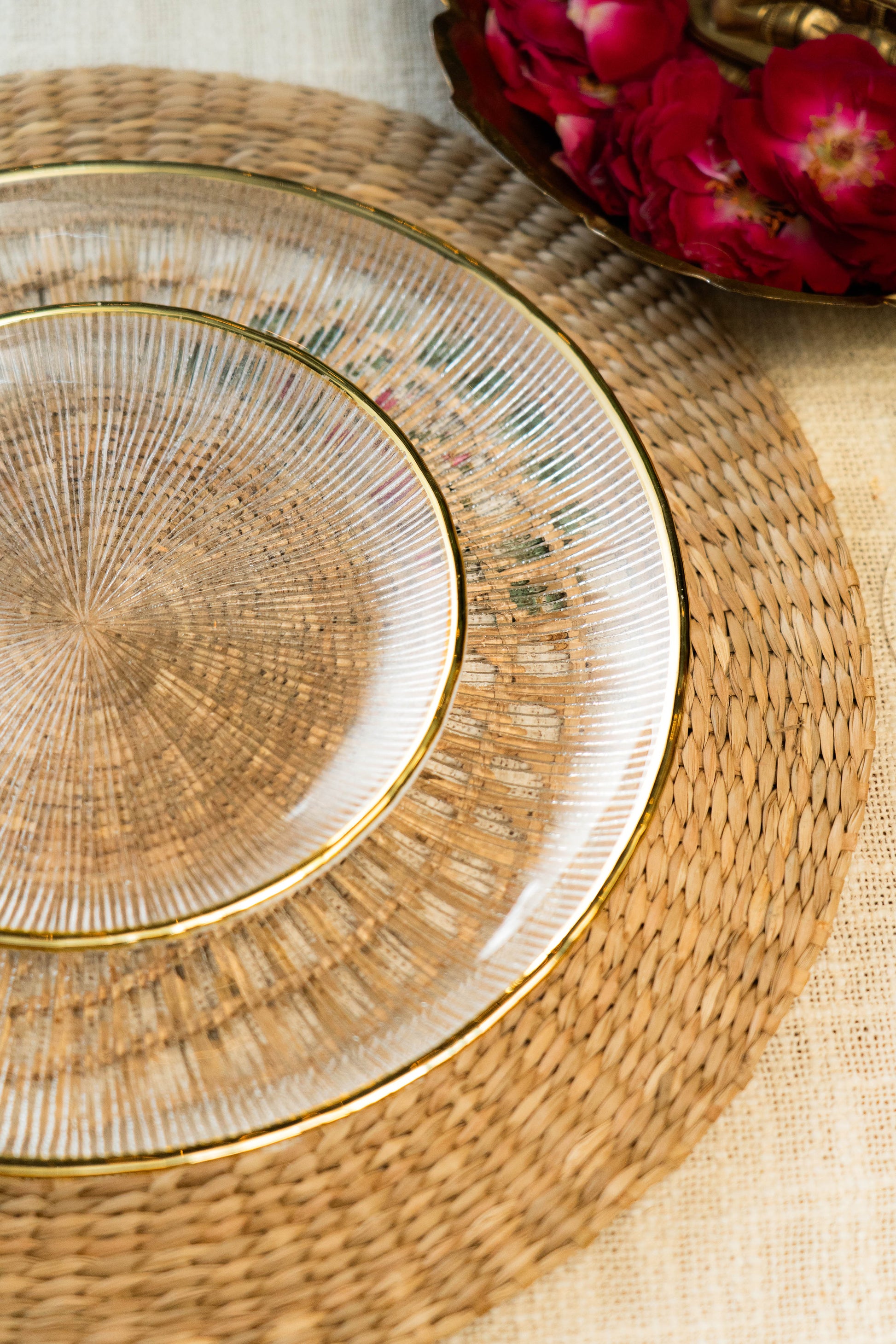 Set of gold-rimmed glass plates on a woven placemat with flowers in the background