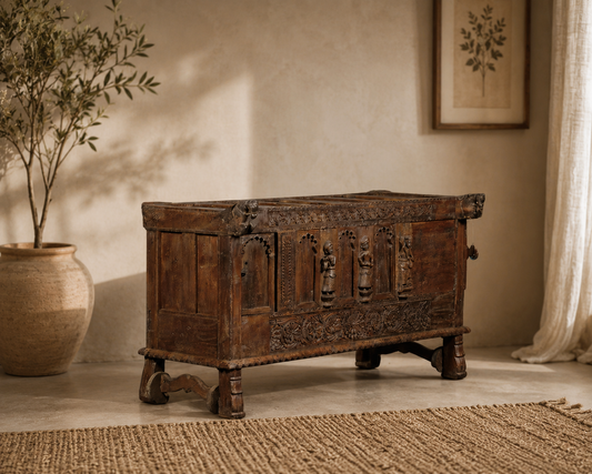 An antique carved wooden chest or trunk with ornate relief details, shown in a styled interior setting with a potted plant, textured rug, and warm wall lighting.