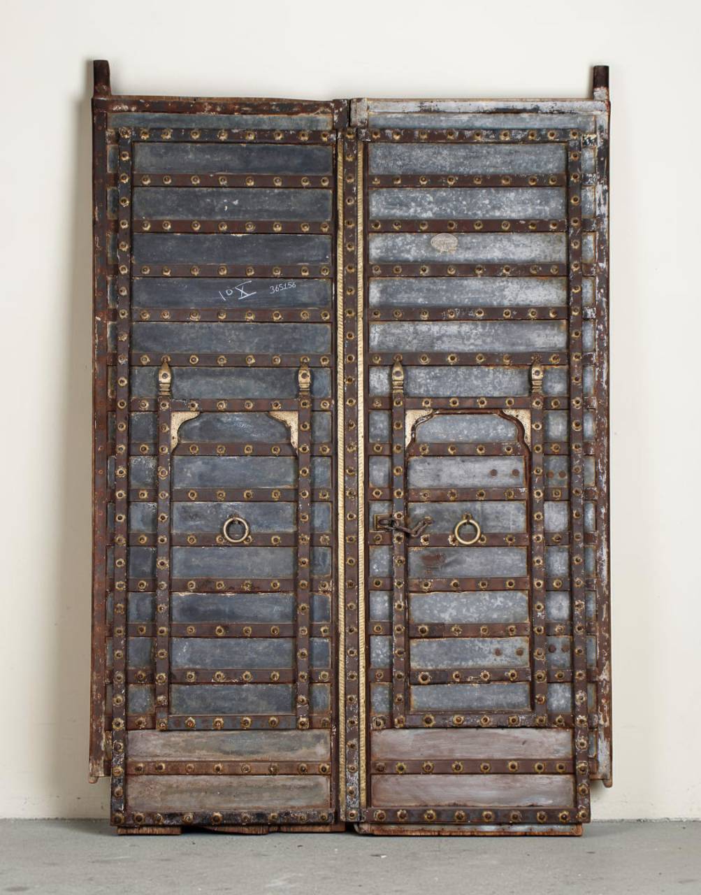 A pair of vintage double doors with weathered gray wood panels, heavy brass rivets, and brass metal loops, standing against a plain off-white wall.