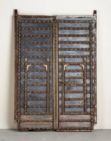 A pair of vintage double doors with weathered gray wood panels, heavy brass rivets, and brass metal loops, standing against a plain off-white wall.