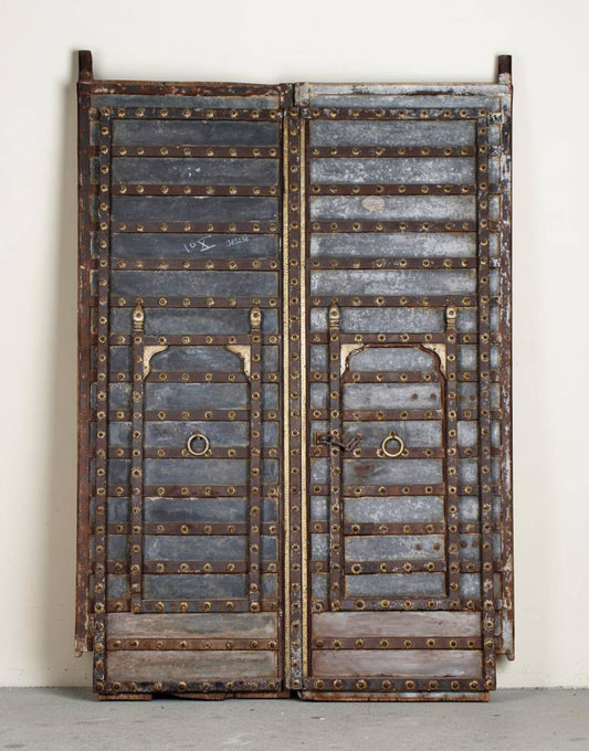A pair of vintage double doors with weathered gray wood panels, heavy brass rivets, and brass metal loops, standing against a plain off-white wall.