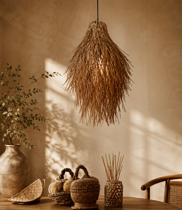 A bohemian-style pendant light made of natural woven reed or straw hangs over a wooden table in a warm, sunlit room. 