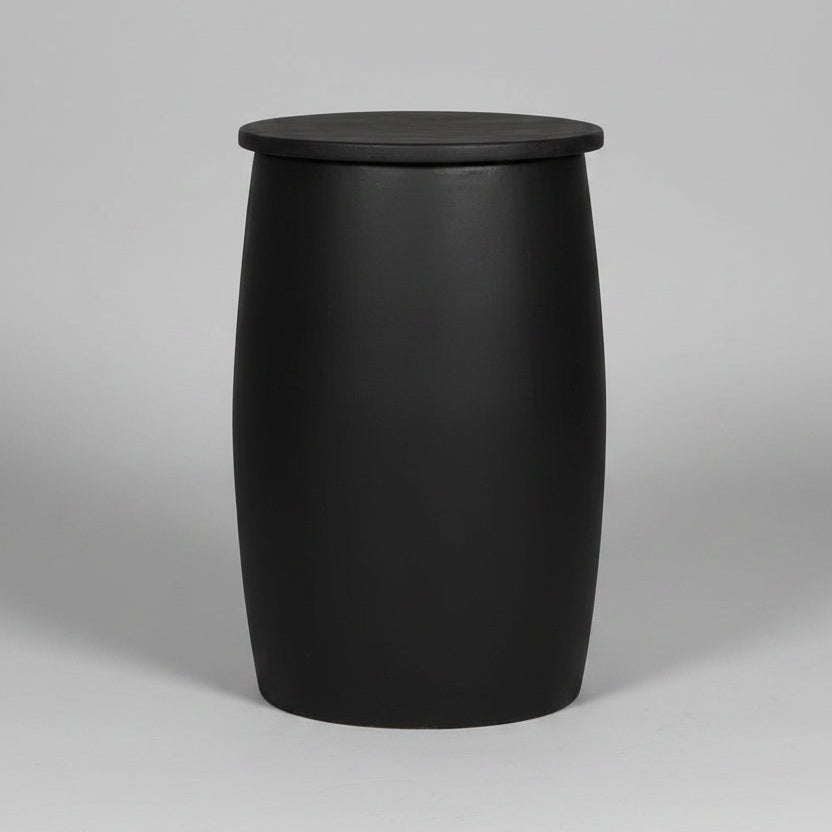 A black barrel-shaped side table or stool with a flat circular top, standing on a light wood floor against a textured gray concrete wall. White handwritten markings are visible on the front.