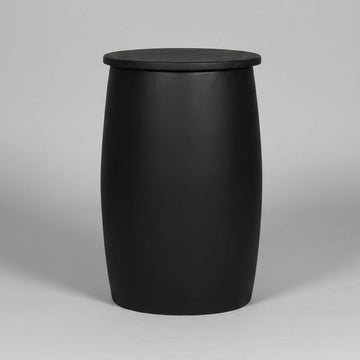 A black barrel-shaped side table or stool with a flat circular top, standing on a light wood floor against a textured gray concrete wall. White handwritten markings are visible on the front.
