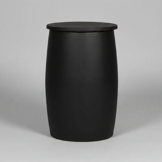 A black barrel-shaped side table or stool with a flat circular top, standing on a light wood floor against a textured gray concrete wall. White handwritten markings are visible on the front.